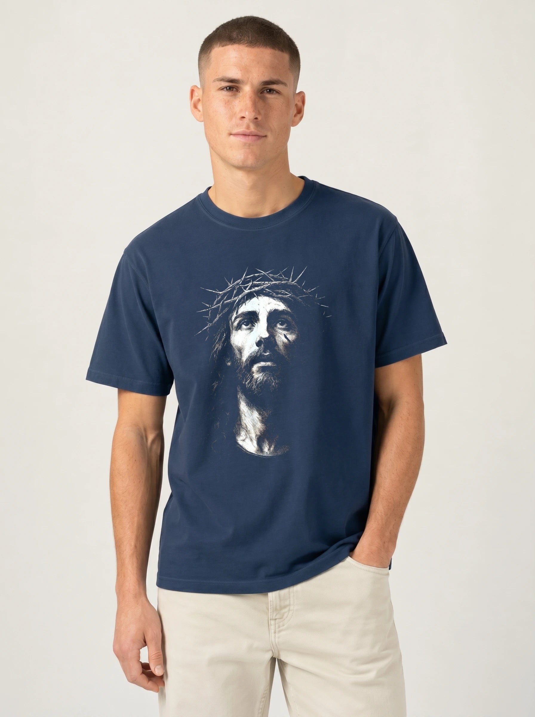 A man wearing a navy blue crewneck t-shirt with a graphic print of Jesus Christ's face on the front, paired with light beige pants, against a plain light gray background.