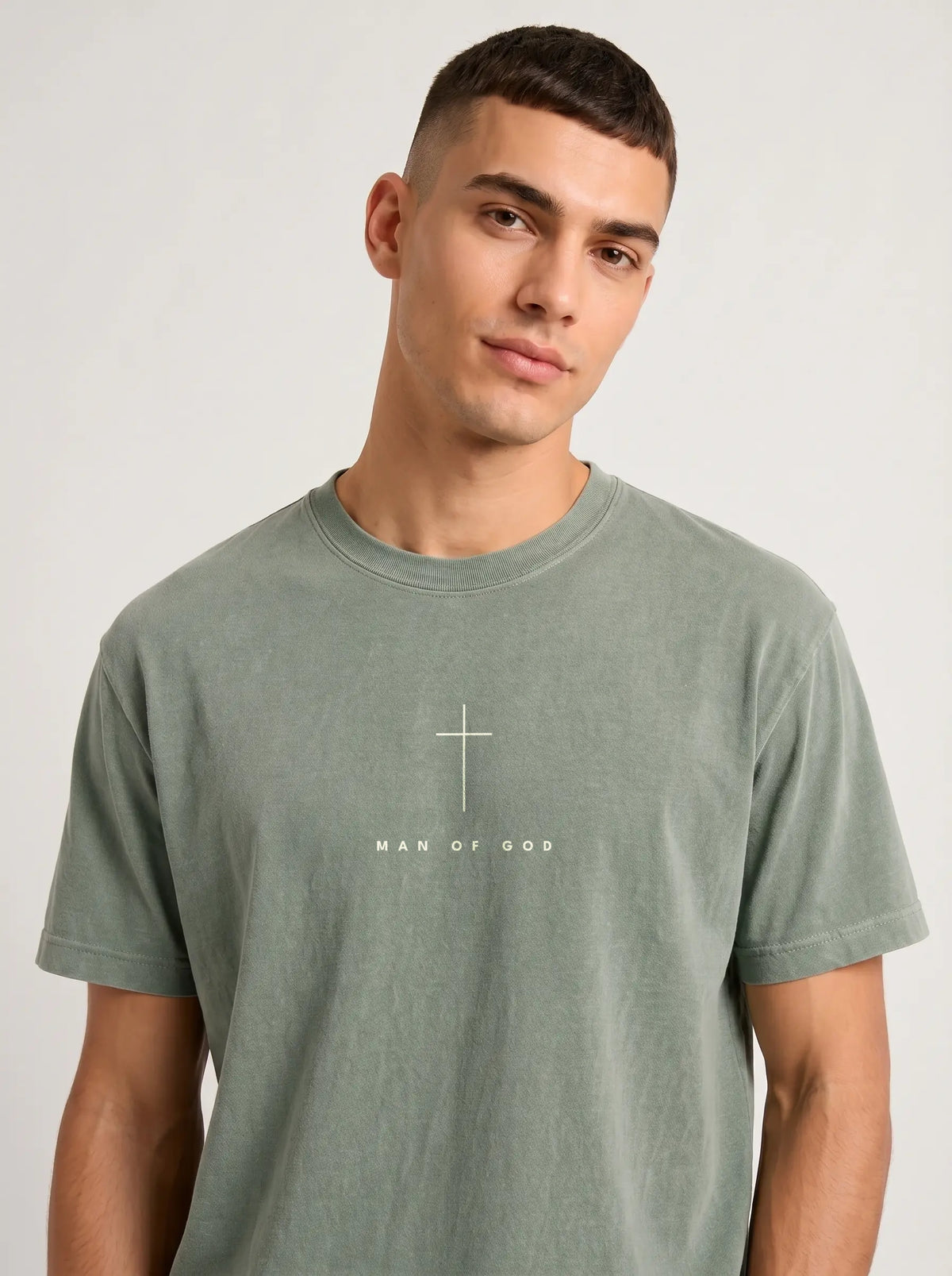 A man wearing a muted sage green t-shirt with 'MAN OF GOD' printed in white text across the chest.
