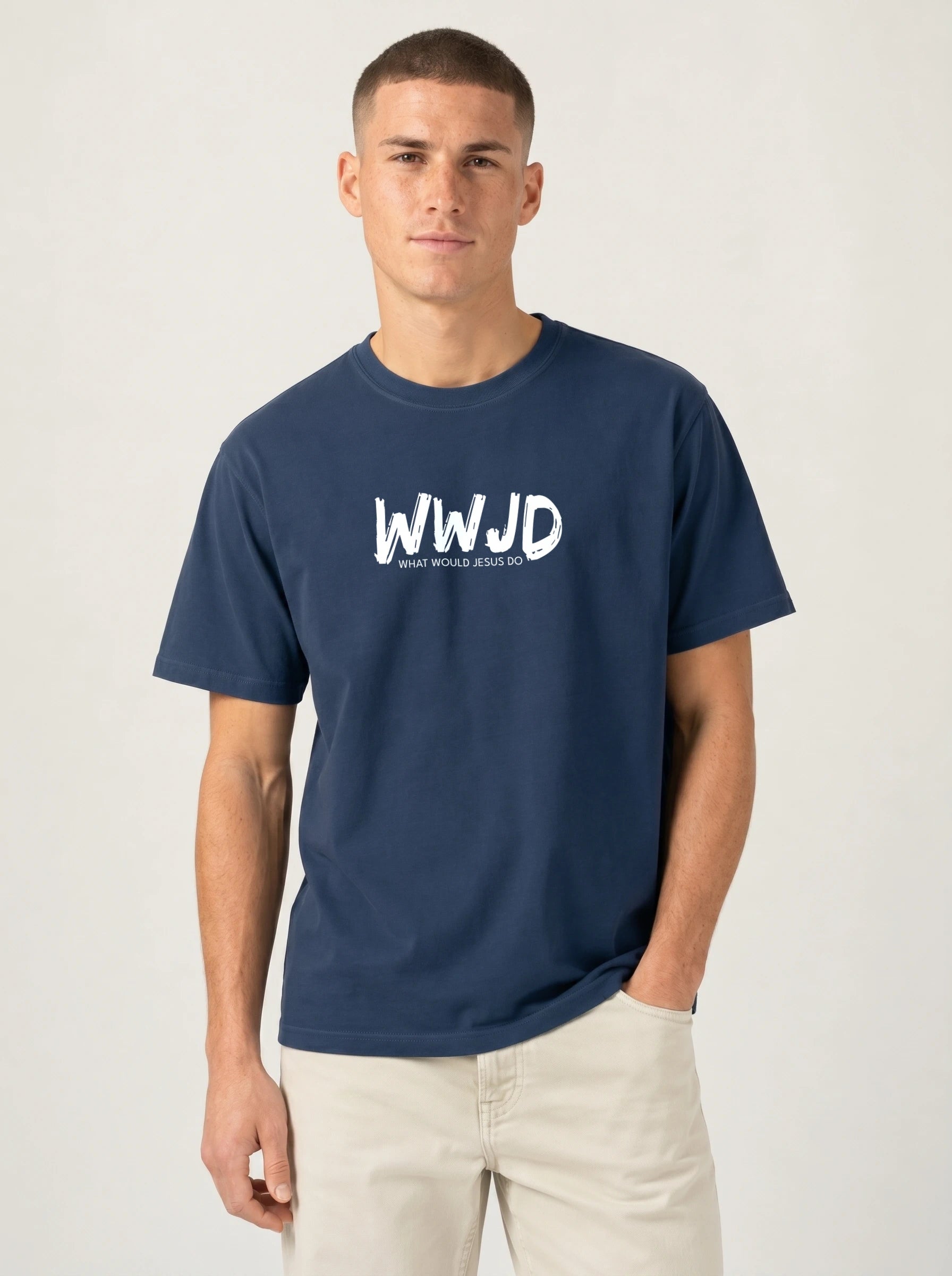 A man wearing a navy blue crewneck t-shirt with a white 'WWJD' graphic on the chest, paired with light beige pants, standing against a light gray studio background.