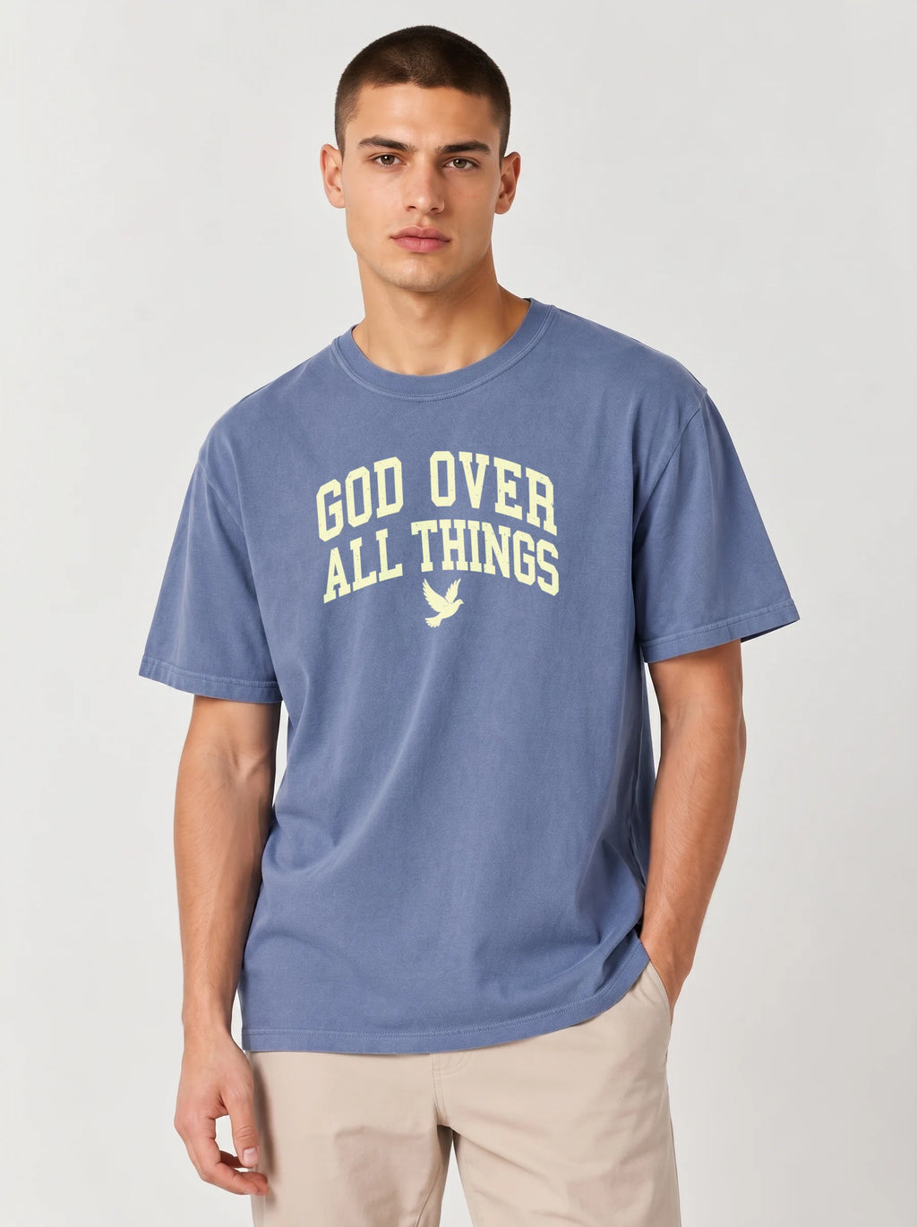 A man wearing a heathered blue t-shirt with 'GOD OVER ALL THINGS' printed in cream across the chest, paired with beige pants, against a light gray studio background.