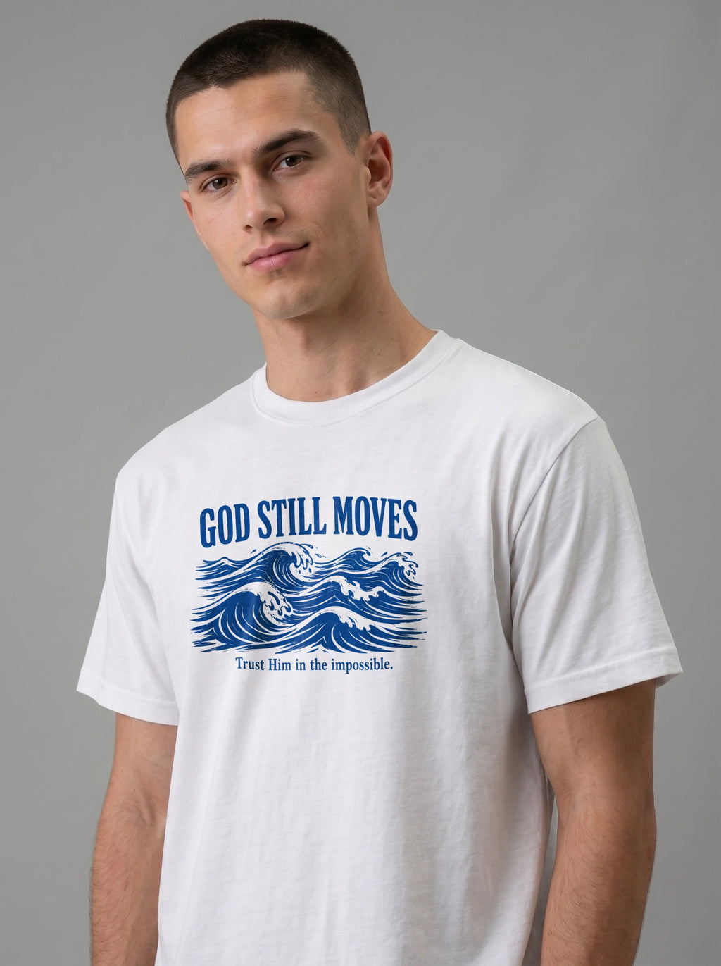 A man wearing a white crewneck t-shirt with a blue graphic print that reads 'GOD STILL MOVES' above blue ocean waves, and 'Trust Him in the impossible.' below, against a solid gray studio background.
