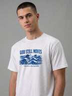 A man wearing a white crewneck t-shirt with a blue graphic print that reads 'GOD STILL MOVES' above blue ocean waves, and 'Trust Him in the impossible.' below, against a solid gray studio background.