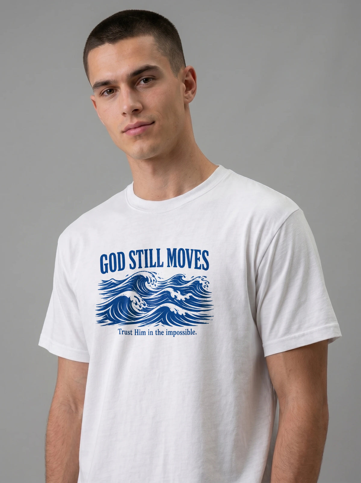 A man wearing a white crewneck t-shirt with a blue graphic print that reads 'GOD STILL MOVES' above blue ocean waves, and 'Trust Him in the impossible.' below, against a solid gray studio background.