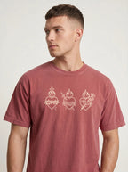 A man wearing a washed dusty rose t-shirt with three white Sacred Heart line drawings on the chest, photographed against a light gray studio background.