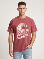 A man wearing a faded red crewneck t-shirt with a graphic of a lion and cross with the text 'MAN OF FAITH' printed on the front, paired with light blue jeans, against a light gray studio background.