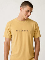 A man wearing a mustard yellow crewneck t-shirt with the word 'REDEEMED' printed in black serif letters across the chest, photographed against a light off-white background.