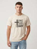 A man wearing a beige crewneck t-shirt with a black graphic print that reads 'I CAN'T BUT I KNOW A GUY' next to a cross, paired with light blue jeans, against a light gray studio background.
