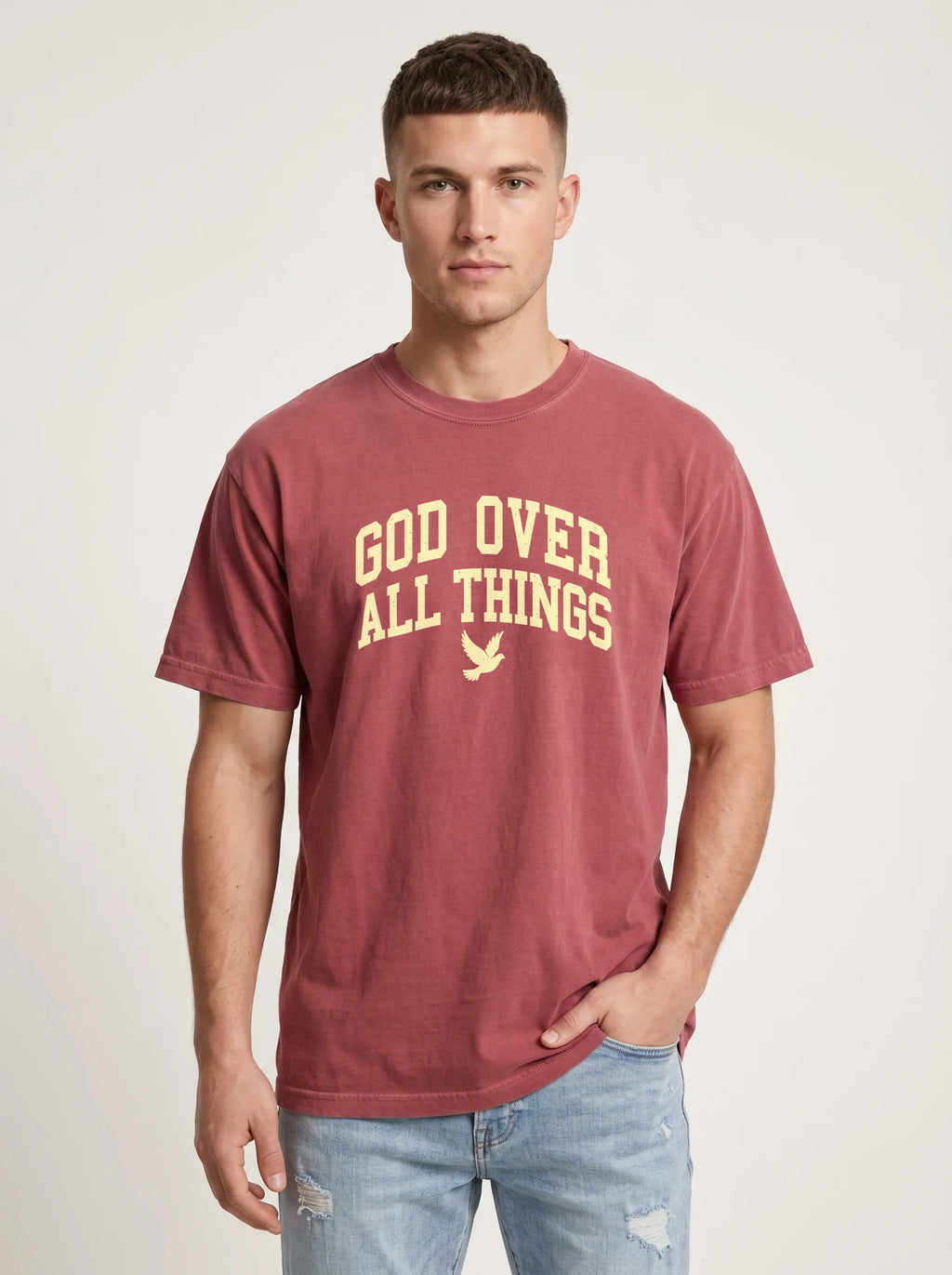A man wearing a faded maroon t-shirt with 'GOD OVER ALL THINGS' printed in yellow on the front, paired with light-wash blue jeans, against a light gray studio background.