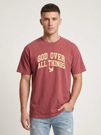 A man wearing a faded maroon t-shirt with 'GOD OVER ALL THINGS' printed in yellow on the front, paired with light-wash blue jeans, against a light gray studio background.