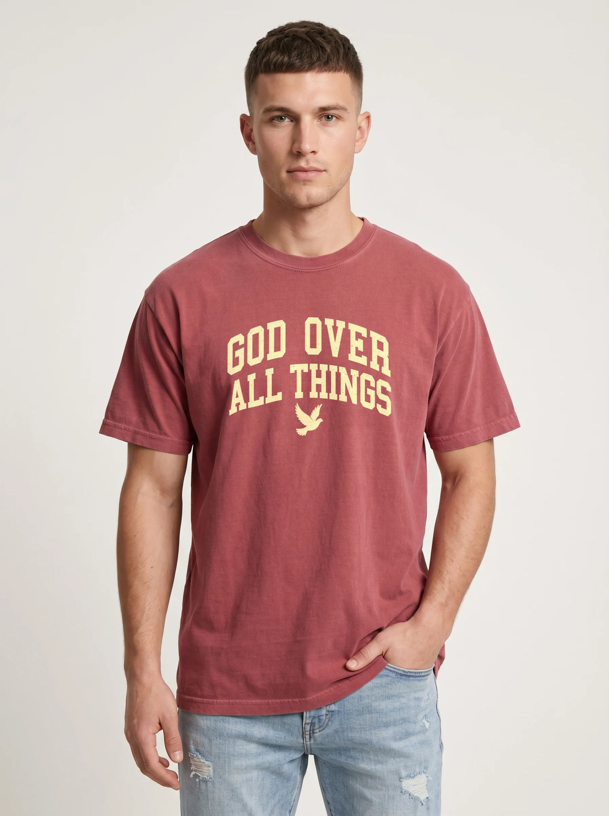 A man wearing a faded maroon t-shirt with 'GOD OVER ALL THINGS' printed in yellow on the front, paired with light-wash blue jeans, against a light gray studio background.