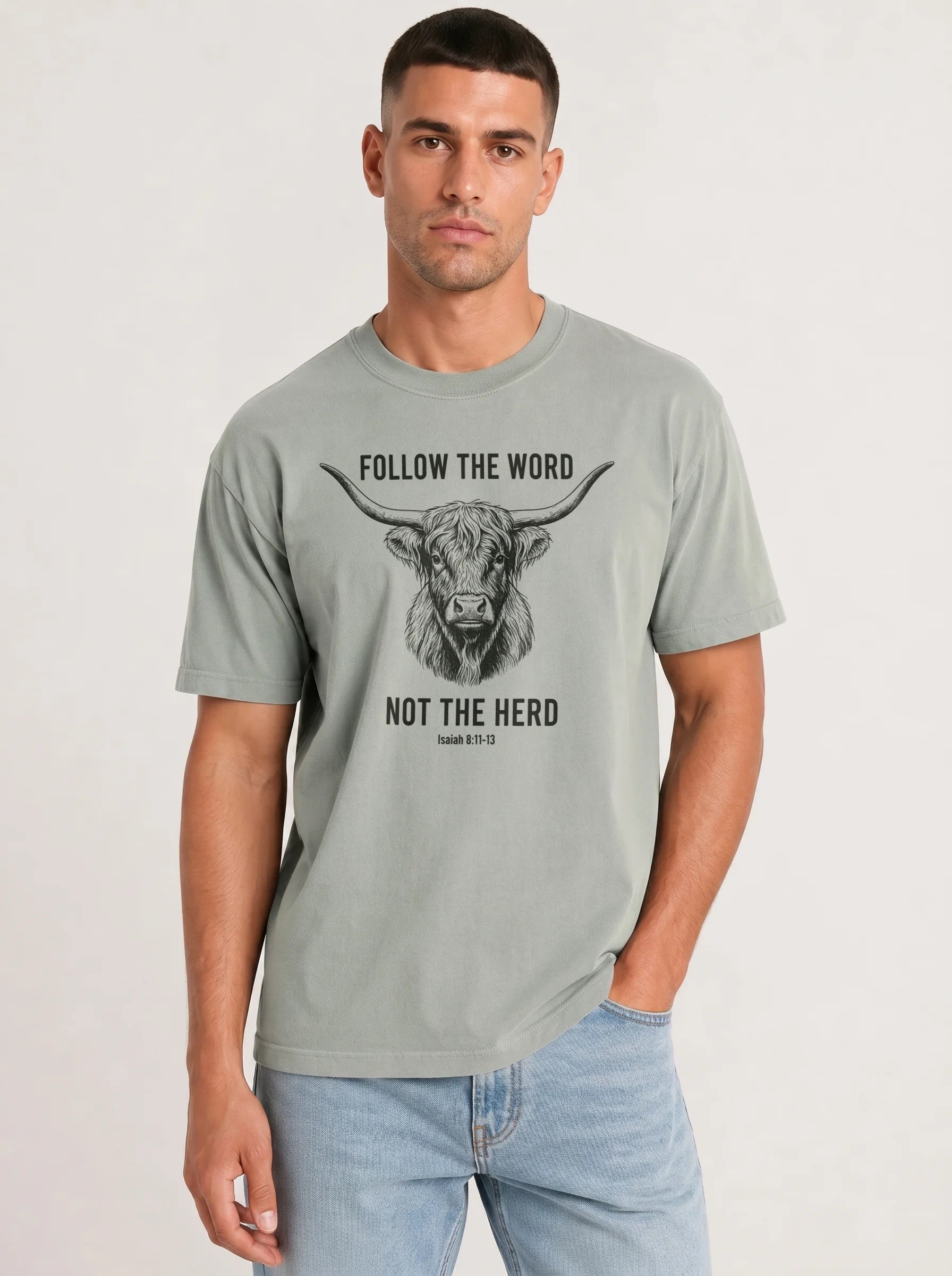 A man wearing a light gray crewneck t-shirt with a black graphic of a highland cow and the text 'FOLLOW THE WORD NOT THE HERD' and 'Isaiah 8:11-13', paired with light blue jeans, against a plain light gray background.