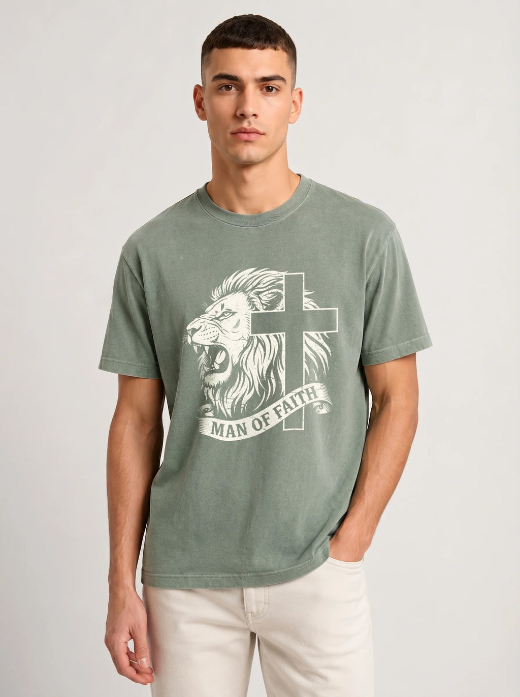 A man wearing a washed sage green t-shirt with a white graphic of a lion and cross with the text 'MAN OF FAITH' on the front, paired with off-white pants, against a light gray studio background.