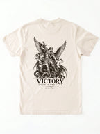 Cream T-shirt laid flat with a winged warrior defeating a dragon and the text “VICTORY OVER DARKNESS” and “REVELATION 12.”