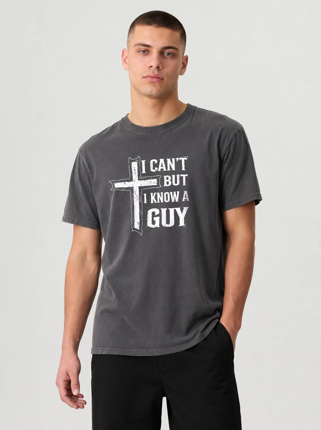 A man wearing a dark gray graphic t-shirt with a white distressed cross and the text 'I CAN'T BUT I KNOW A GUY' printed on the chest, paired with black pants, against a light gray studio background.