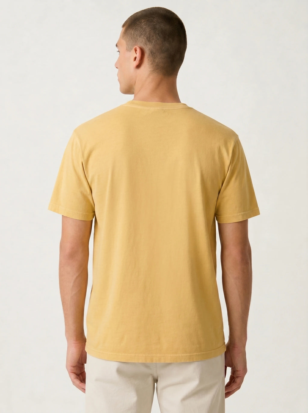 A man viewed from the back wearing a plain short-sleeve crew-neck t-shirt in a warm mustard yellow color, paired with off-white trousers, against a light gray studio background.