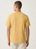 A man viewed from the back wearing a plain short-sleeve crew-neck t-shirt in a warm mustard yellow color, paired with off-white trousers, against a light gray studio background.