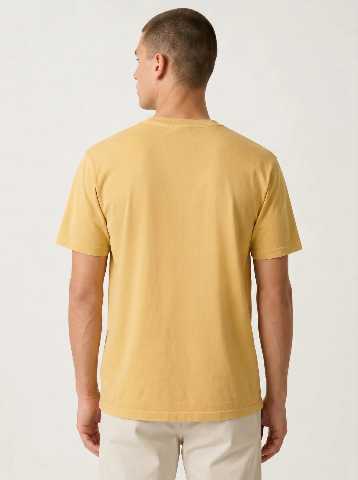 A man viewed from the back wearing a plain short-sleeve crew-neck t-shirt in a warm mustard yellow color, paired with off-white trousers, against a light gray studio background.