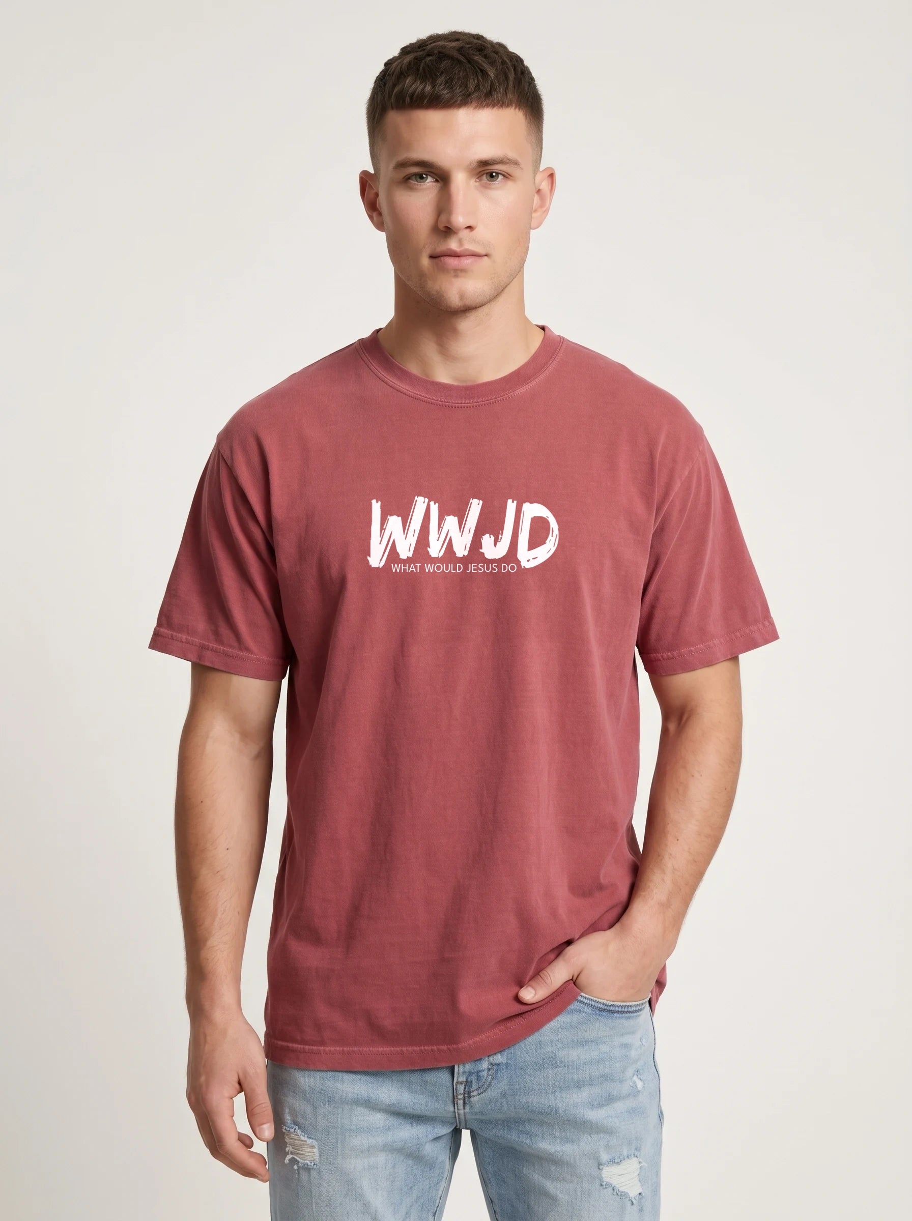A man wearing a washed red crewneck t-shirt with a white 'WWJD' graphic on the chest, paired with light blue jeans, standing against a light gray studio background.