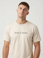 A man wearing a light beige crewneck t-shirt with the text 'JESUS + SAVES' printed in black across the chest, set against a plain light gray background.