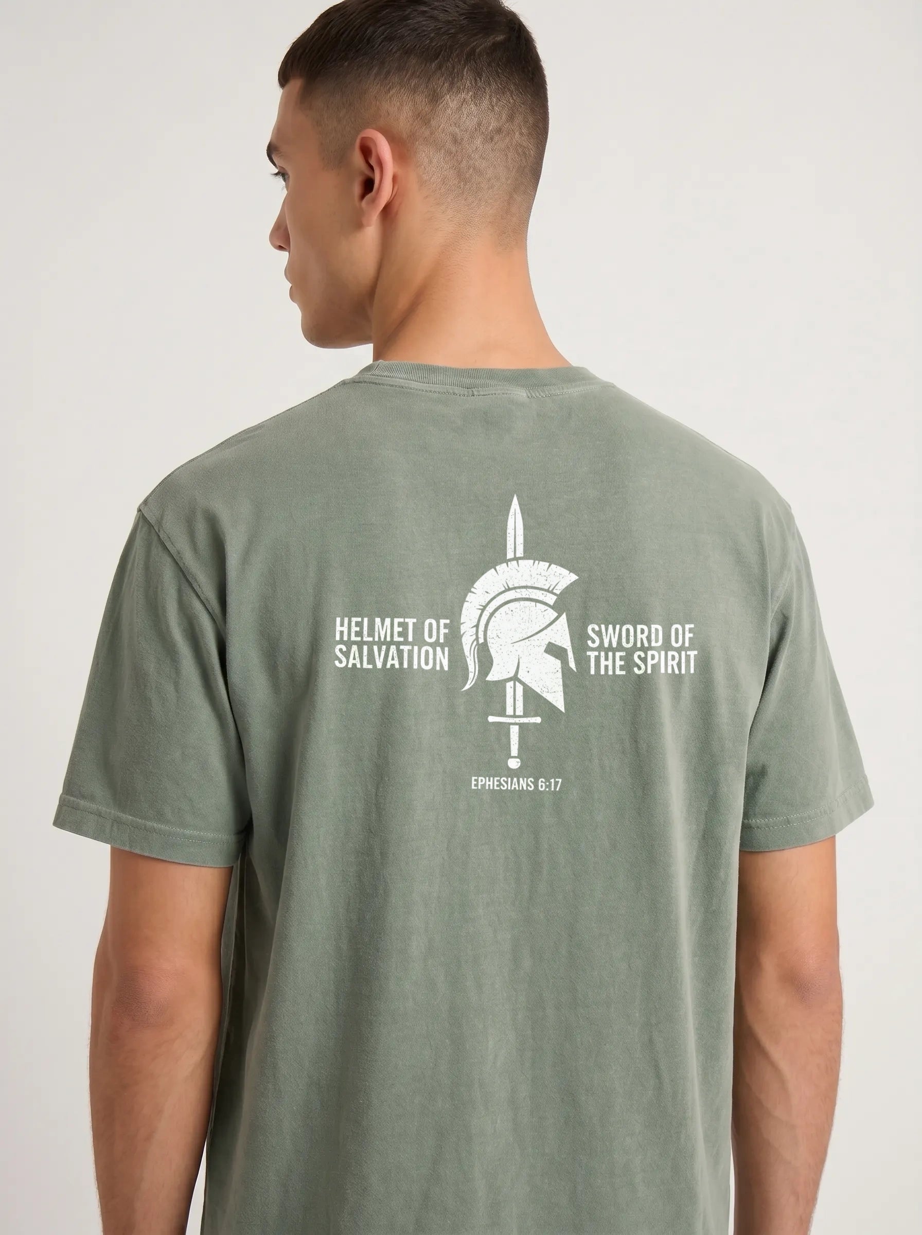 A man wearing a sage green t-shirt, viewed from the back. The shirt features a white graphic print of a Spartan helmet and sword with the text 'HELMET OF SALVATION' and 'SWORD OF THE SPIRIT' and 'ESPHANSAS 6:17'.