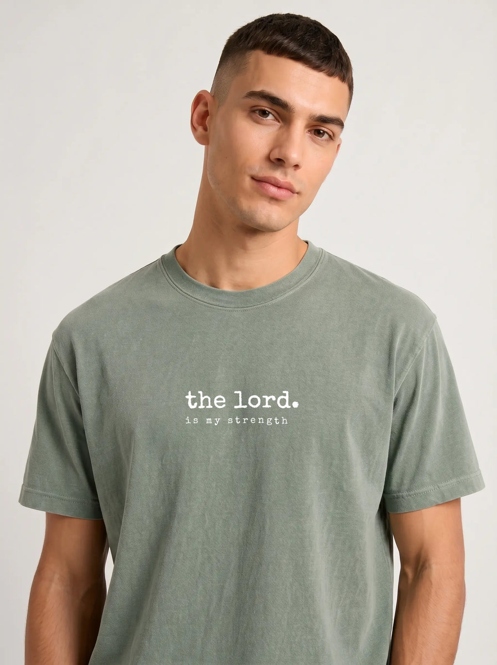 A man wearing a muted sage green crewneck t-shirt with the white text 'the lord. is my strength' printed across the chest, photographed against a neutral light gray background.