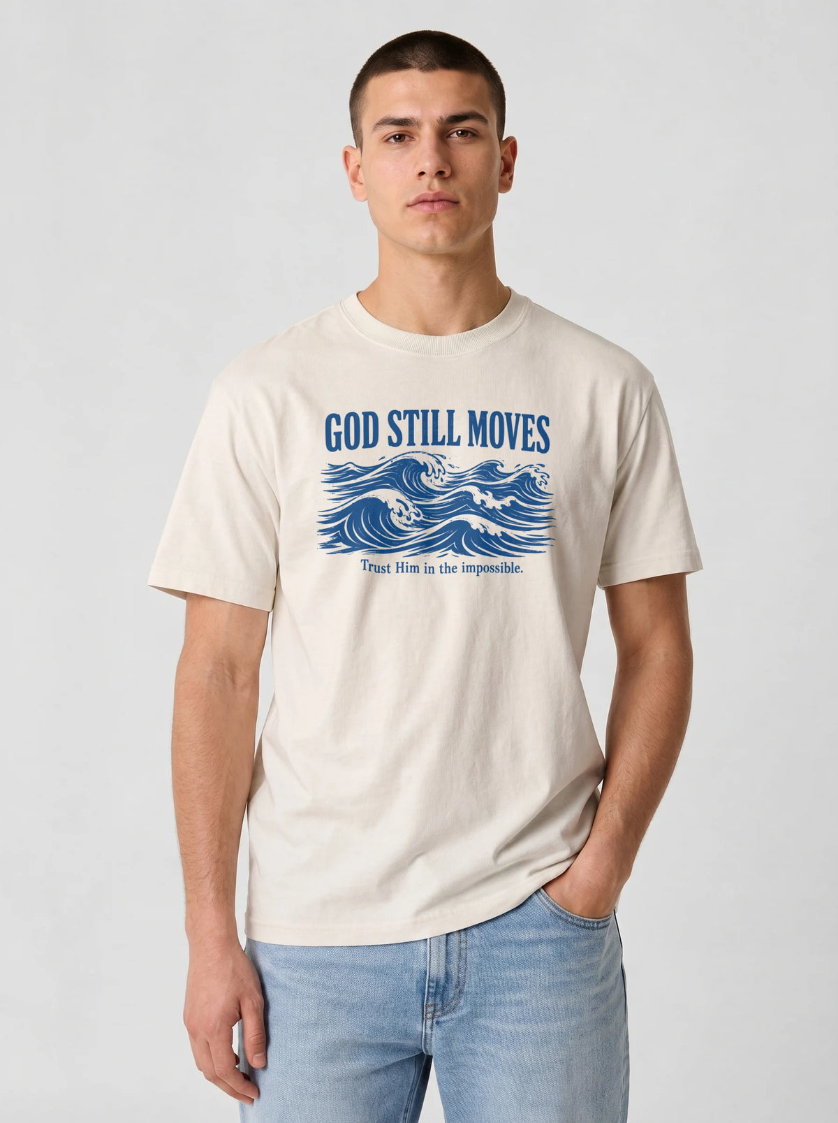 Man wearing a cream Comfort Colors t-shirt with a navy blue wave illustration and the text "GOD STILL MOVES" above "Trust Him in the impossible," paired with light blue jeans against a plain gray studio background.