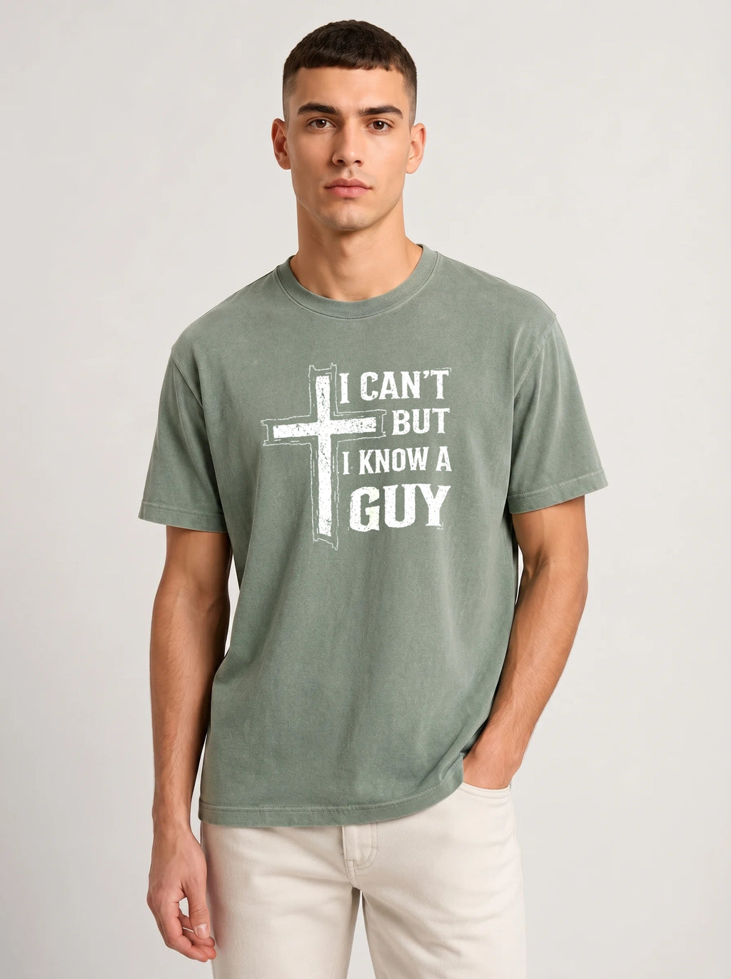 A man wearing a heather sage green t-shirt with a white graphic print that reads 'I CAN'T BUT I KNOW A GUY', paired with off-white pants, against a light gray studio background.