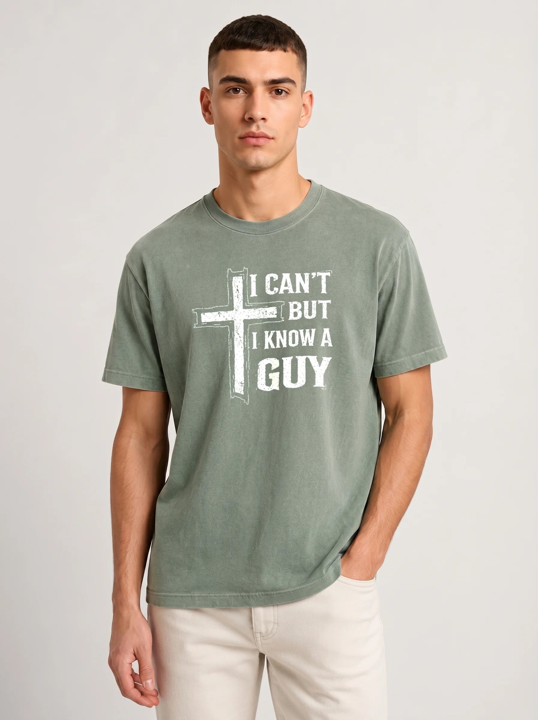 A man wearing a heather sage green t-shirt with a white graphic print that reads 'I CAN'T BUT I KNOW A GUY', paired with off-white pants, against a light gray studio background.