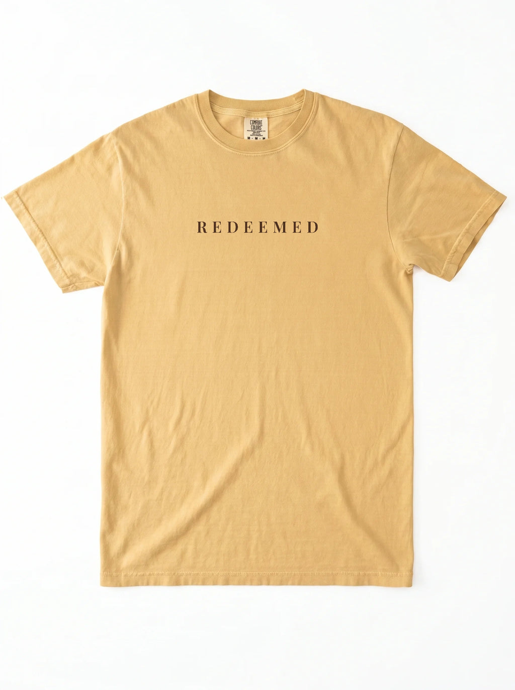 A light tan crewneck t-shirt with the word 'REDEEMED' printed in black serif letters across the chest, displayed flat against a white background.