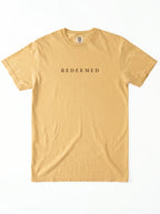 A light tan crewneck t-shirt with the word 'REDEEMED' printed in black serif letters across the chest, displayed flat against a white background.
