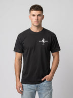 A man wearing a black short-sleeve crewneck t-shirt with a small white graphic on the left chest, paired with light blue jeans, standing against a solid gray studio background.