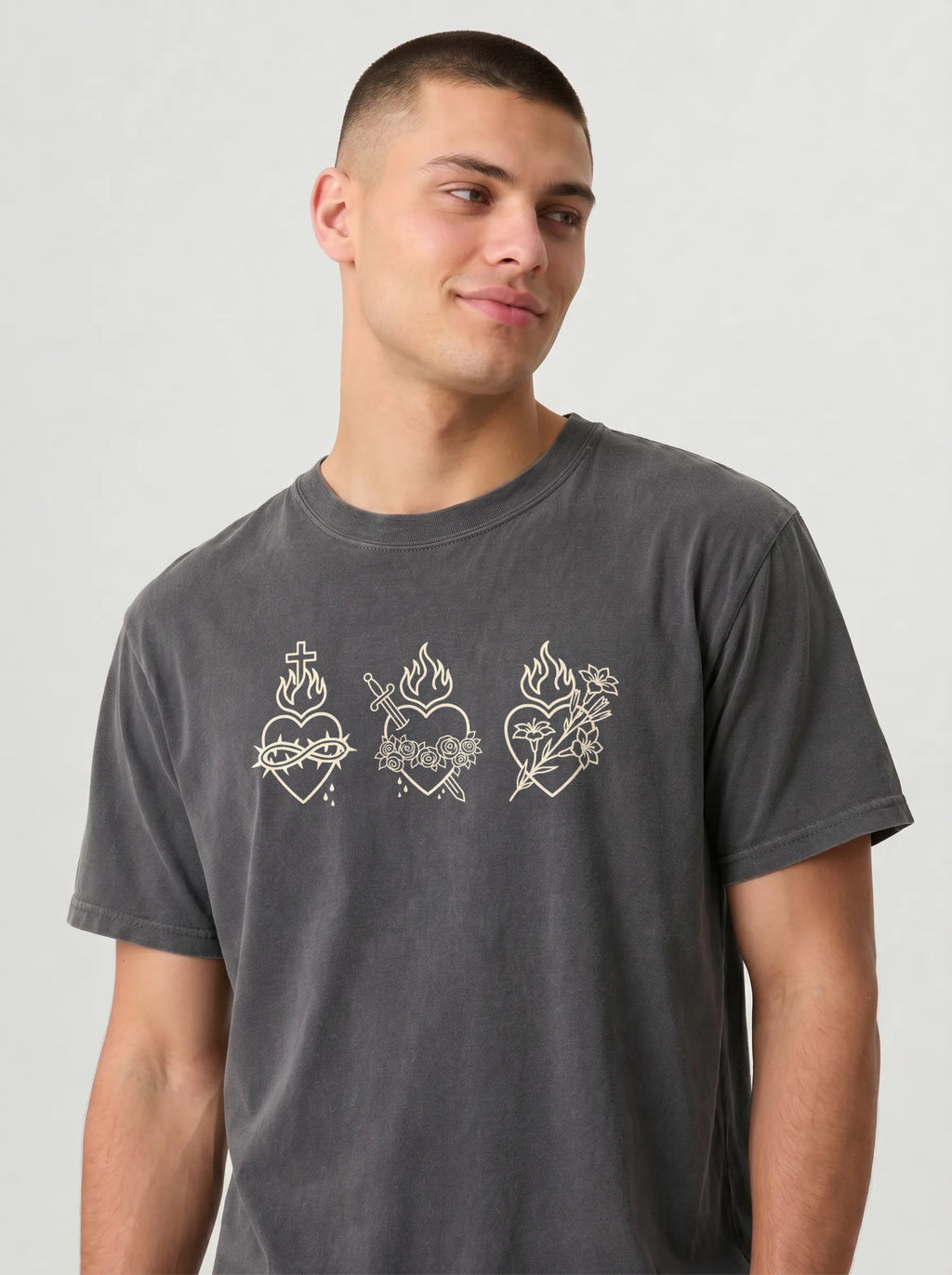 A man wearing a dark gray washed t-shirt with three white line-art sacred heart designs across the chest, photographed against a light gray studio background.
