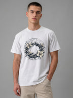 A man wearing a white crewneck t-shirt with a graphic print of a crown of thorns and white lilies, featuring Greek text. He is also wearing beige pants, standing against a solid gray studio background.
