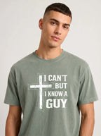 A man wearing a faded sage green t-shirt with a white graphic print that reads 'I CAN'T BUT I KNOW A GUY', set against a plain light gray background.