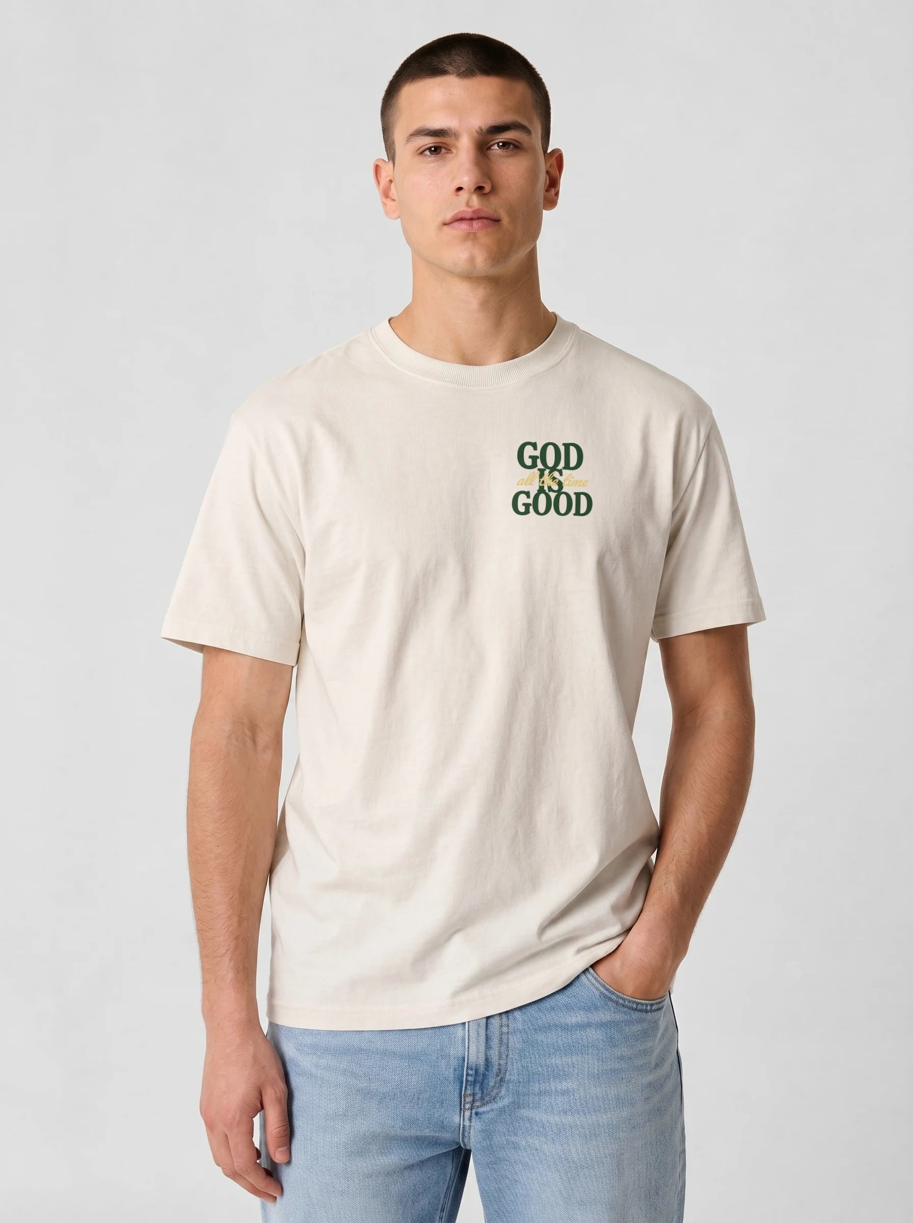 A man wearing a cream-colored t-shirt with 'GOD IS GOOD' printed in green on the chest, paired with light blue jeans, against a light gray studio background.