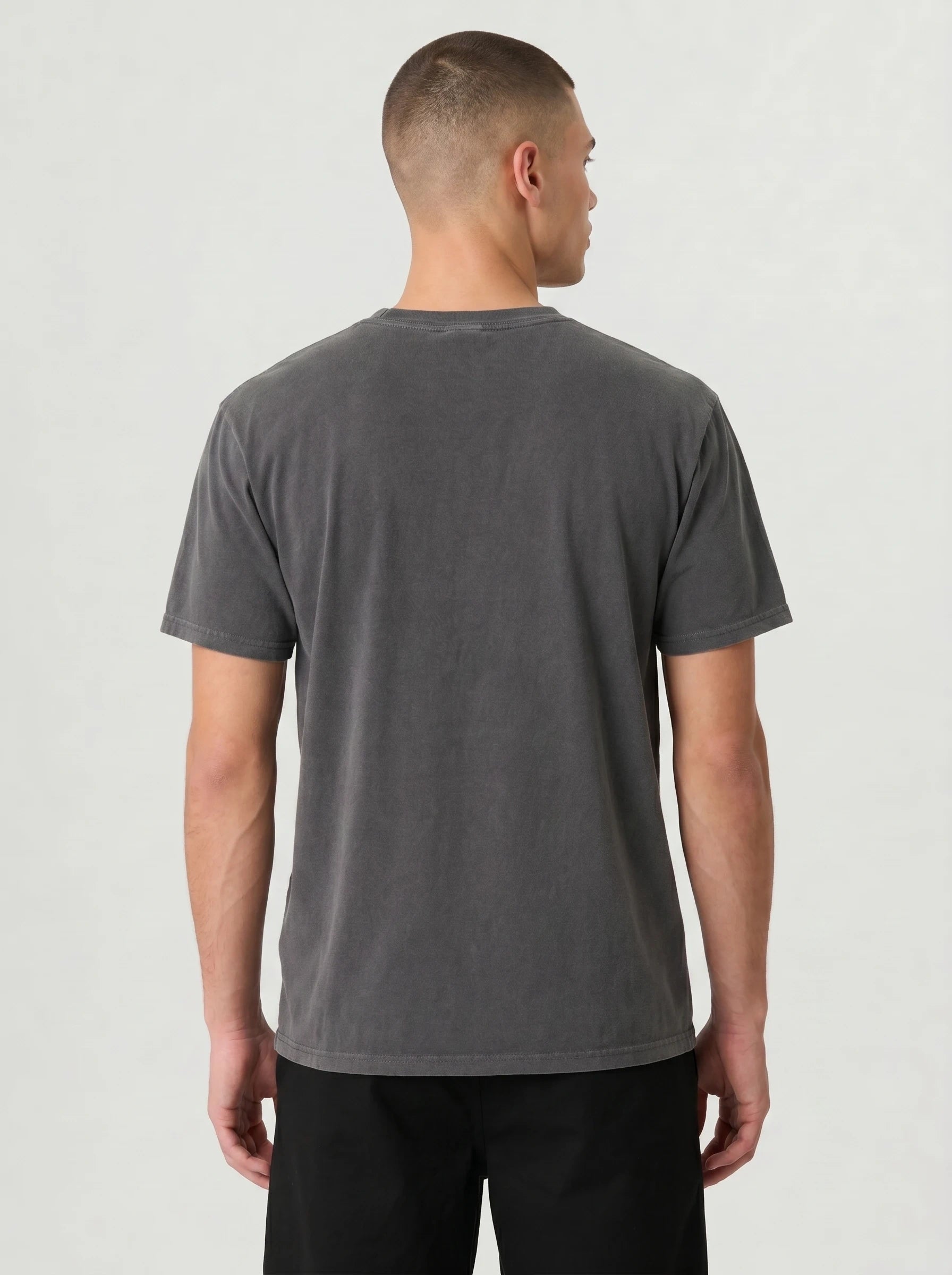 A man viewed from the back wearing a dark gray washed short-sleeve t-shirt and black pants, standing against a light gray studio background.