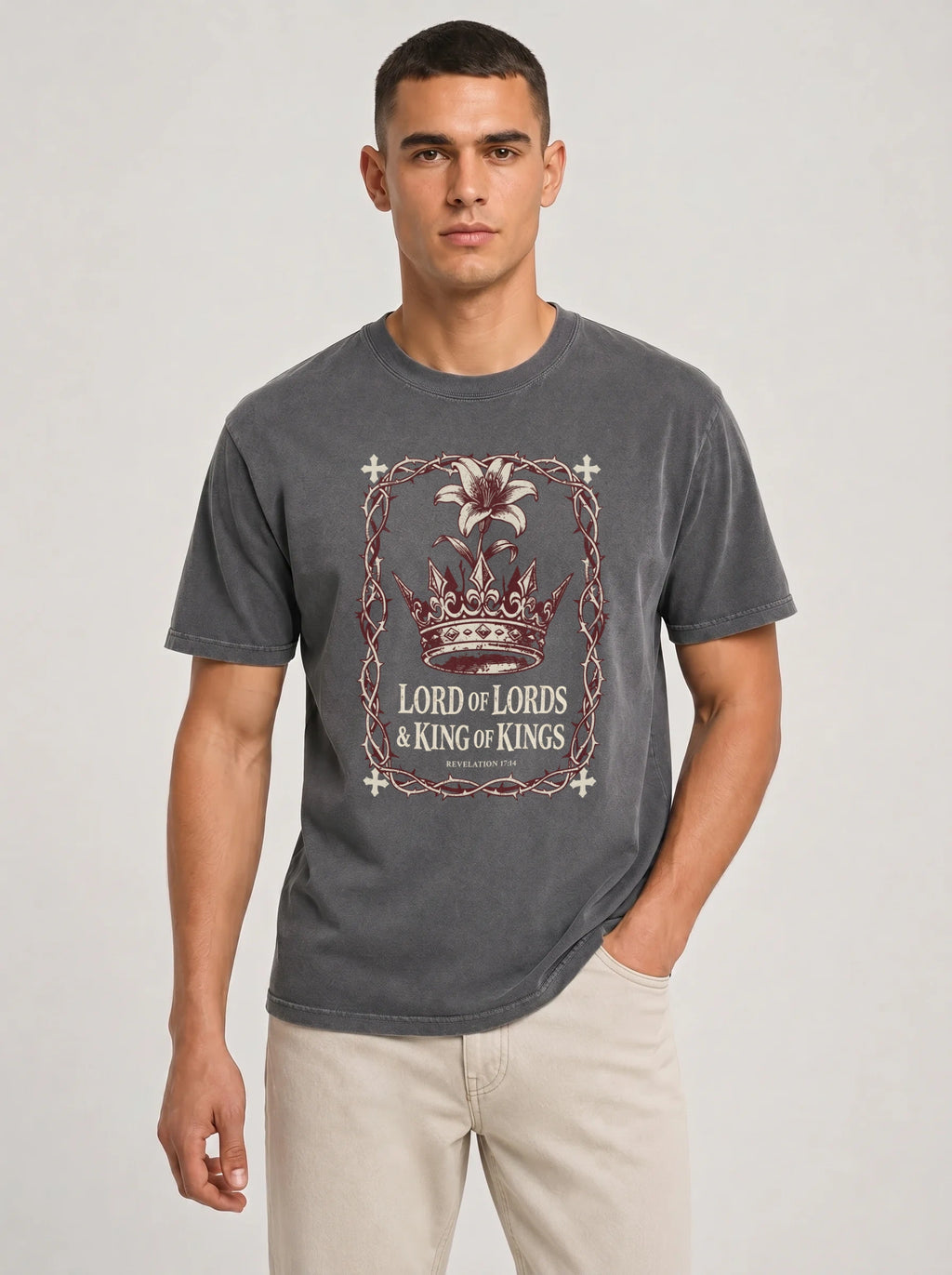 A man wearing a dark gray graphic t-shirt with a crown and 'Lord of Lords & King of Kings' design, paired with beige pants, against a light gray studio background.