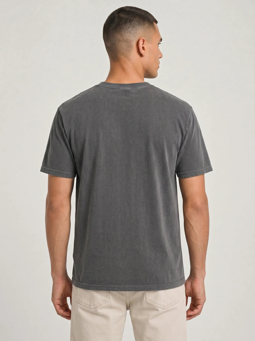A man viewed from the back wearing a dark gray washed short-sleeve t-shirt and light beige pants, set against a plain light gray studio background.