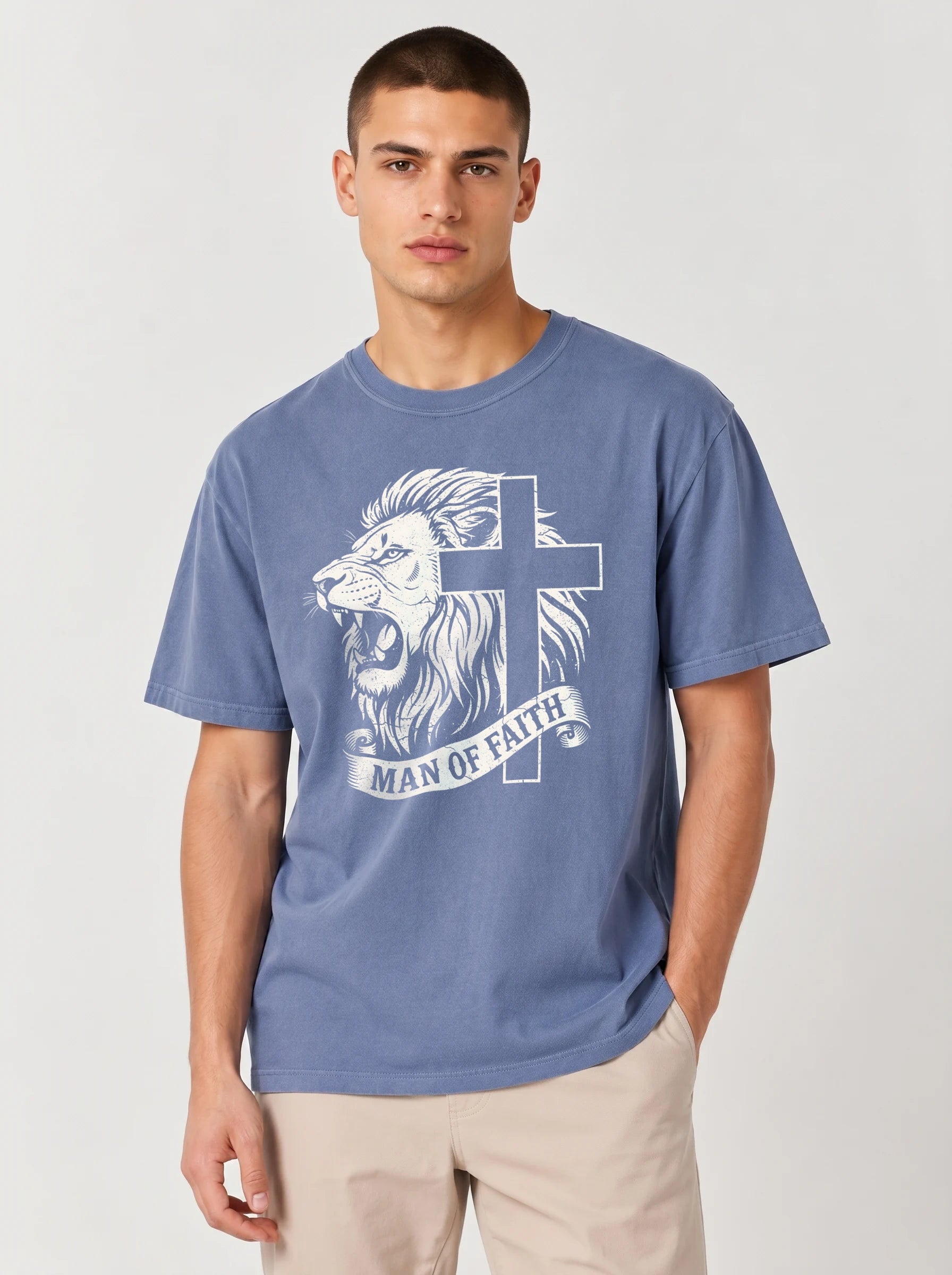 A man wearing a medium blue crewneck t-shirt with a white graphic of a lion head, a cross, and a banner reading 'MAN OF FAITH', paired with beige pants, against a light gray studio background.