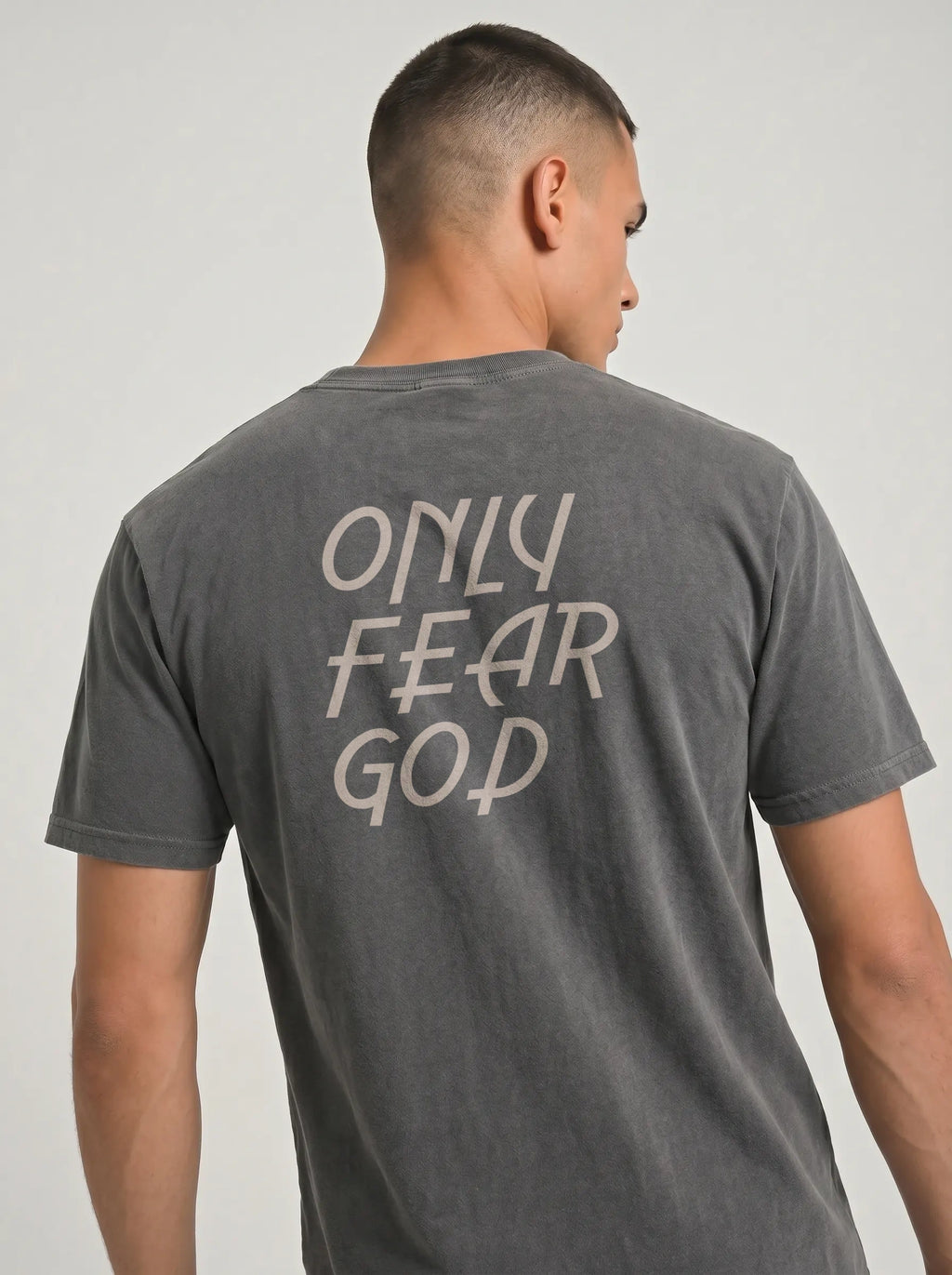 A man wearing a dark gray washed t-shirt, viewed from the back. The shirt features the phrase 'ONLY FEAR GOD' printed in a light beige hand-drawn font across the upper back.