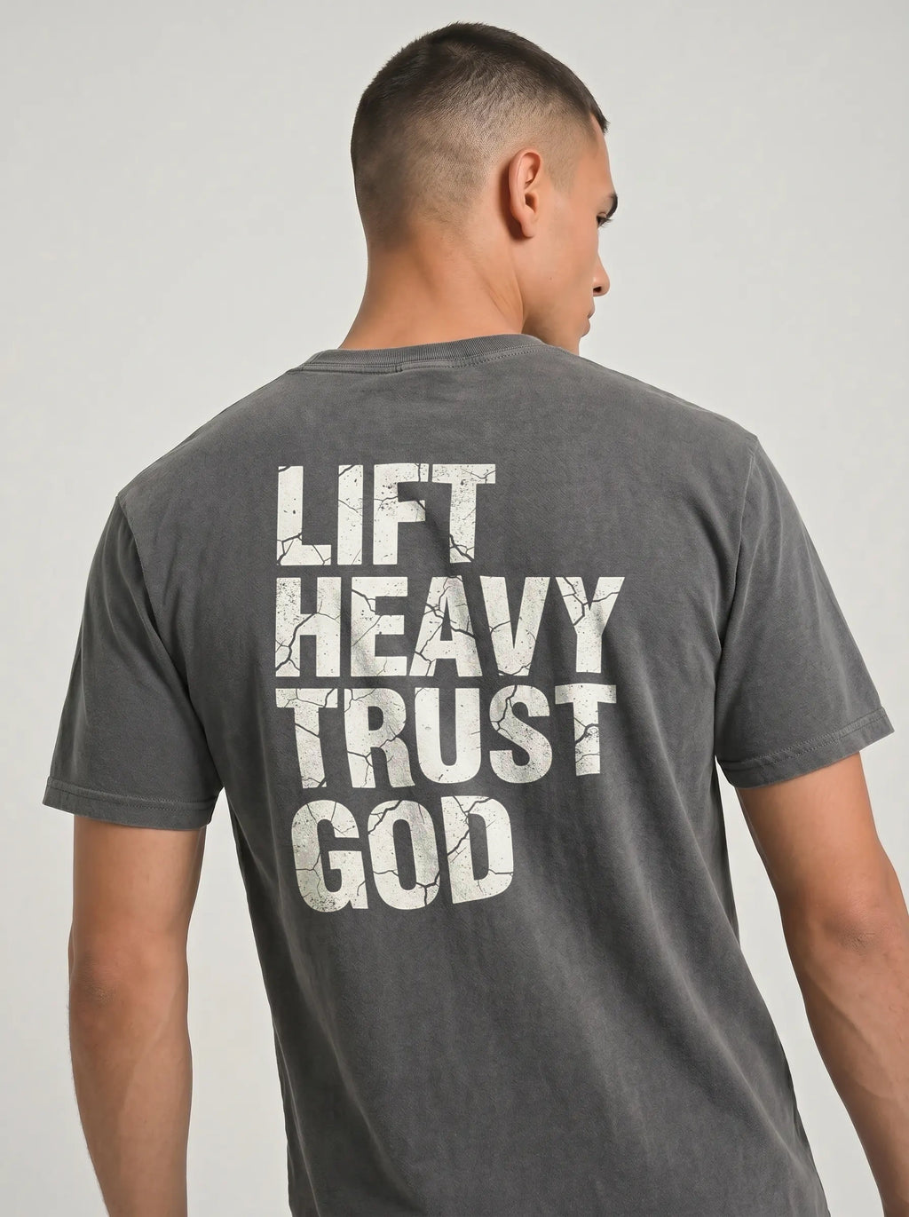 A man viewed from the back wearing a dark gray mineral-wash t-shirt with a distressed white graphic text print that reads 'LIFT HEAVY TRUST GOD'.