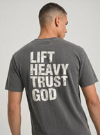 A man viewed from the back wearing a dark gray mineral-wash t-shirt with a distressed white graphic text print that reads 'LIFT HEAVY TRUST GOD'.