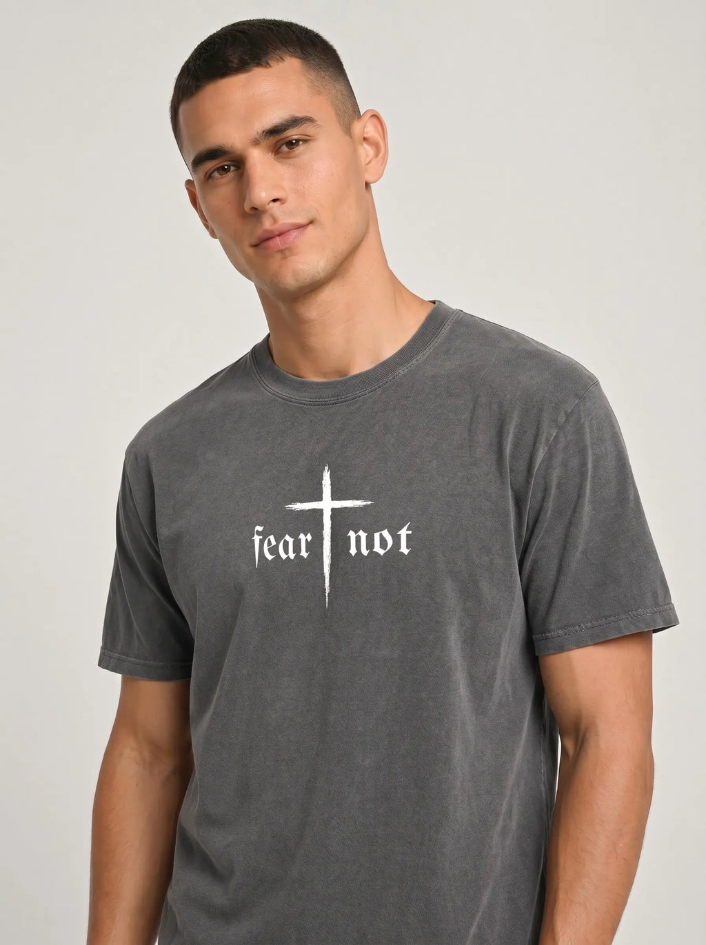 A man wearing a dark gray washed t-shirt with a white graphic print that reads 'fear not' beneath a cross, set against a plain light gray background.