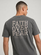 A man wearing a dark gray washed t-shirt, viewed from the back. The shirt features a large distressed light gray graphic on the back reading 'FAITH OVER FEAR' with a smaller biblical quote underneath.