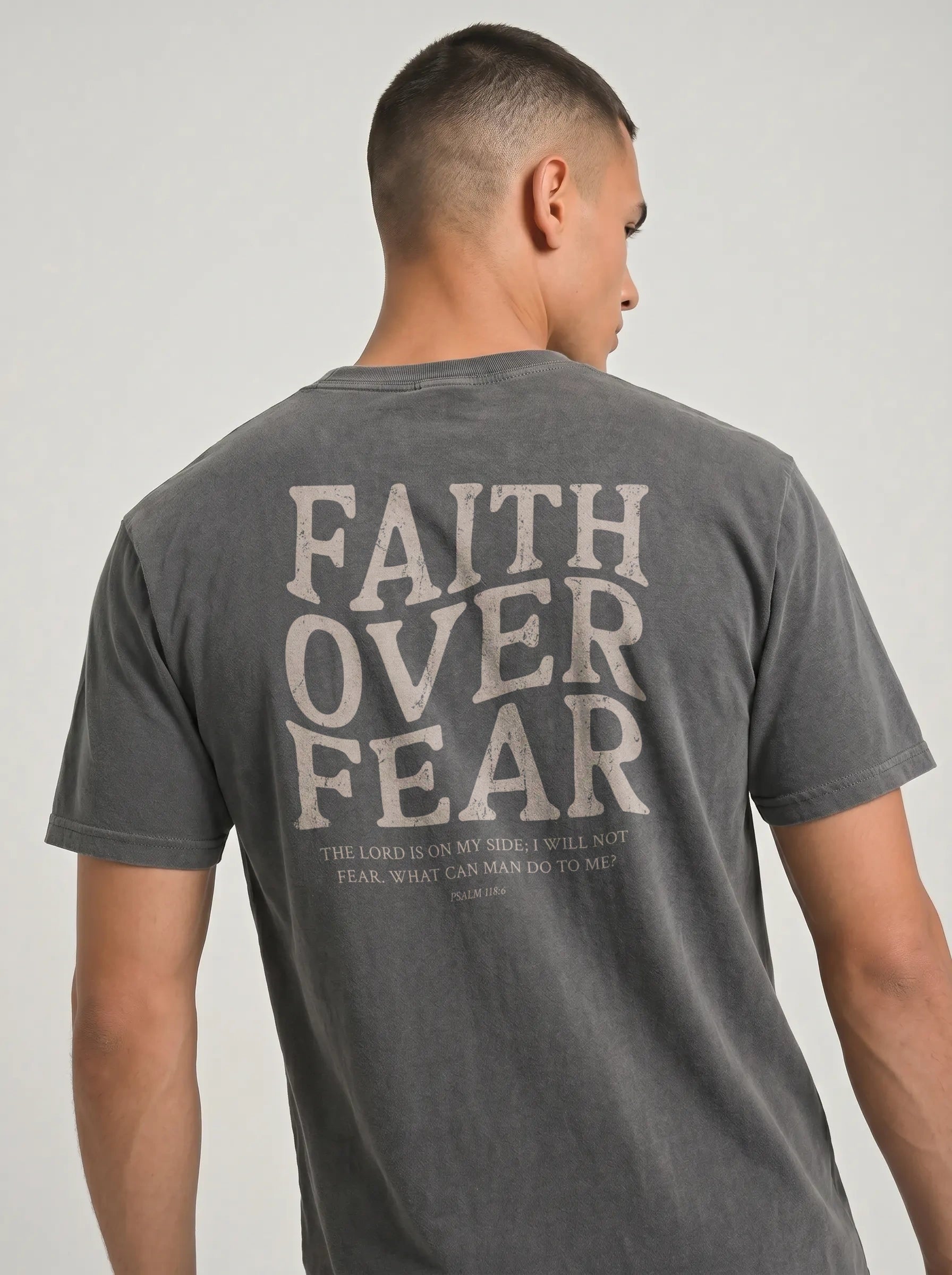 A man wearing a dark gray washed t-shirt, viewed from the back. The shirt features a large distressed light gray graphic on the back reading 'FAITH OVER FEAR' with a smaller biblical quote underneath.