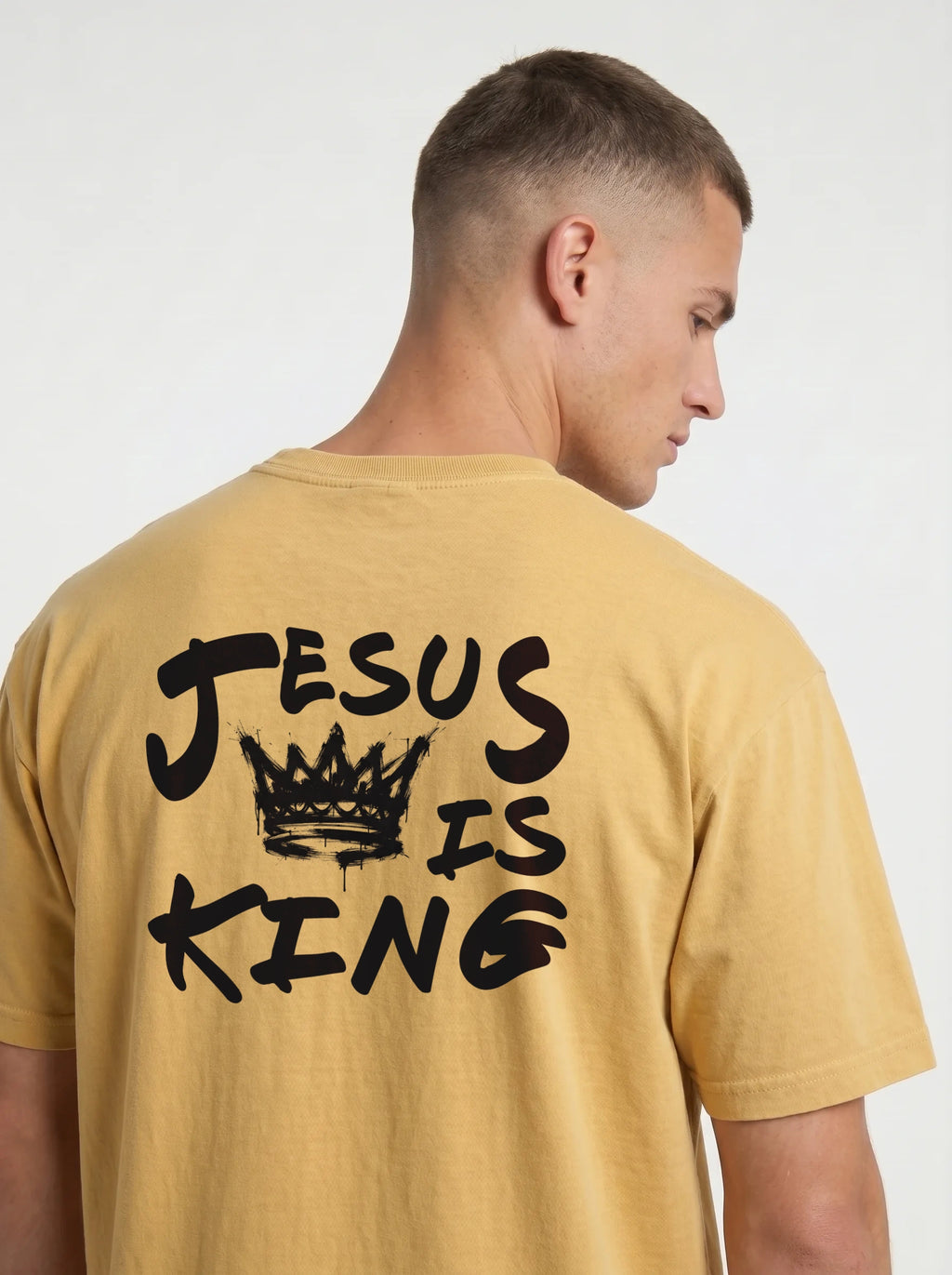 A man wearing a mustard yellow t-shirt from the back, featuring a black graphic print that reads 'JESUS IS KING' with a crown illustration.