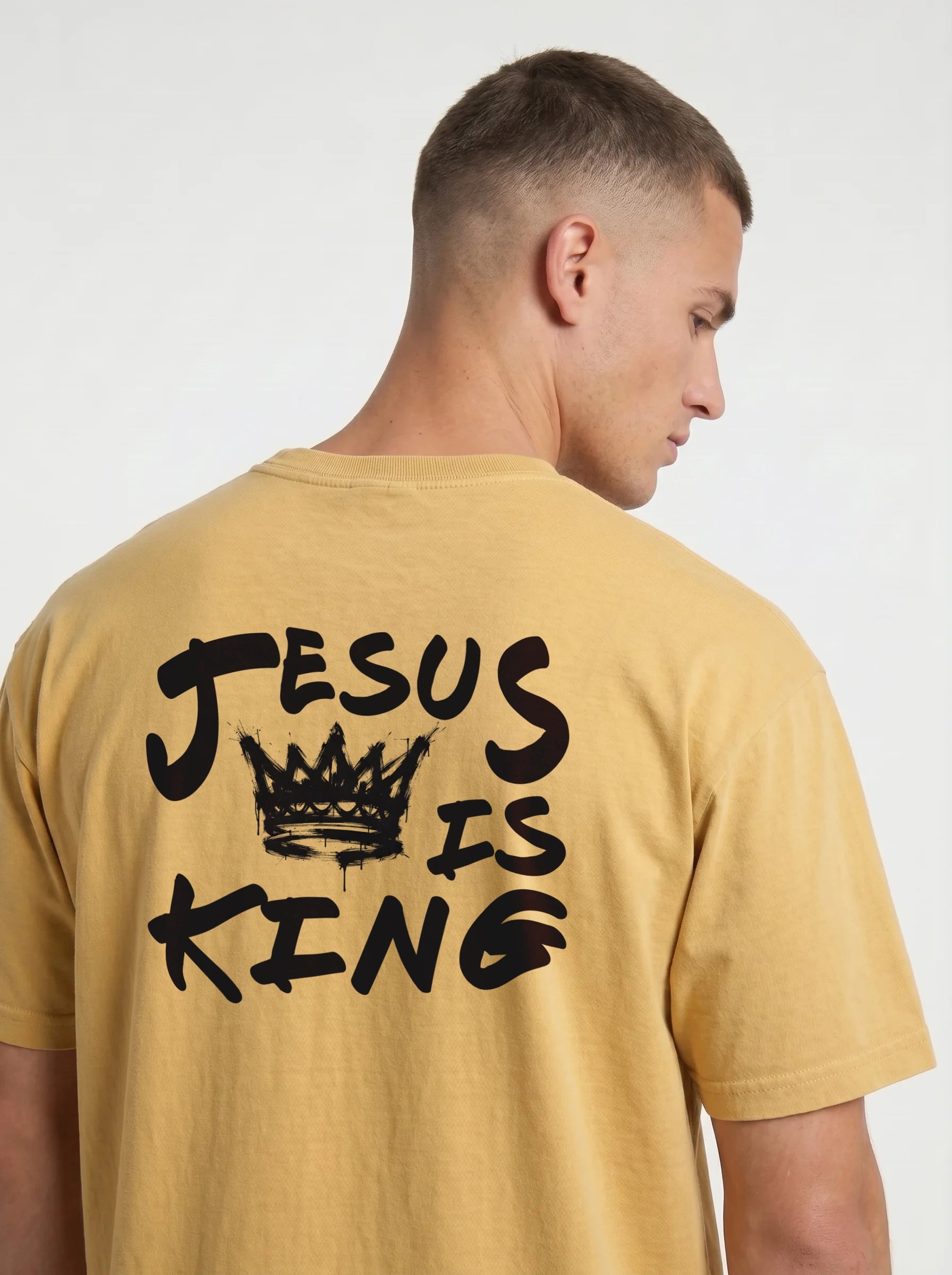 A man wearing a mustard yellow t-shirt from the back, featuring a black graphic print that reads 'JESUS IS KING' with a crown illustration.