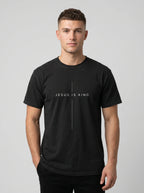 A man wearing a black crewneck t-shirt with a white graphic print that reads 'JESUS IS KING' flanking a vertical cross, photographed against a neutral gray background.