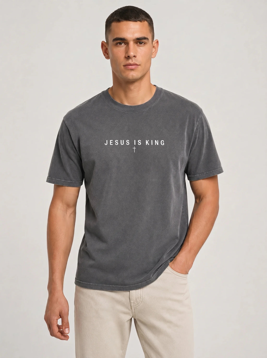 A man wearing a dark gray washed t-shirt with the text 'JESUS IS KING' and a small cross printed on the chest, paired with light beige pants, against a light gray studio background.