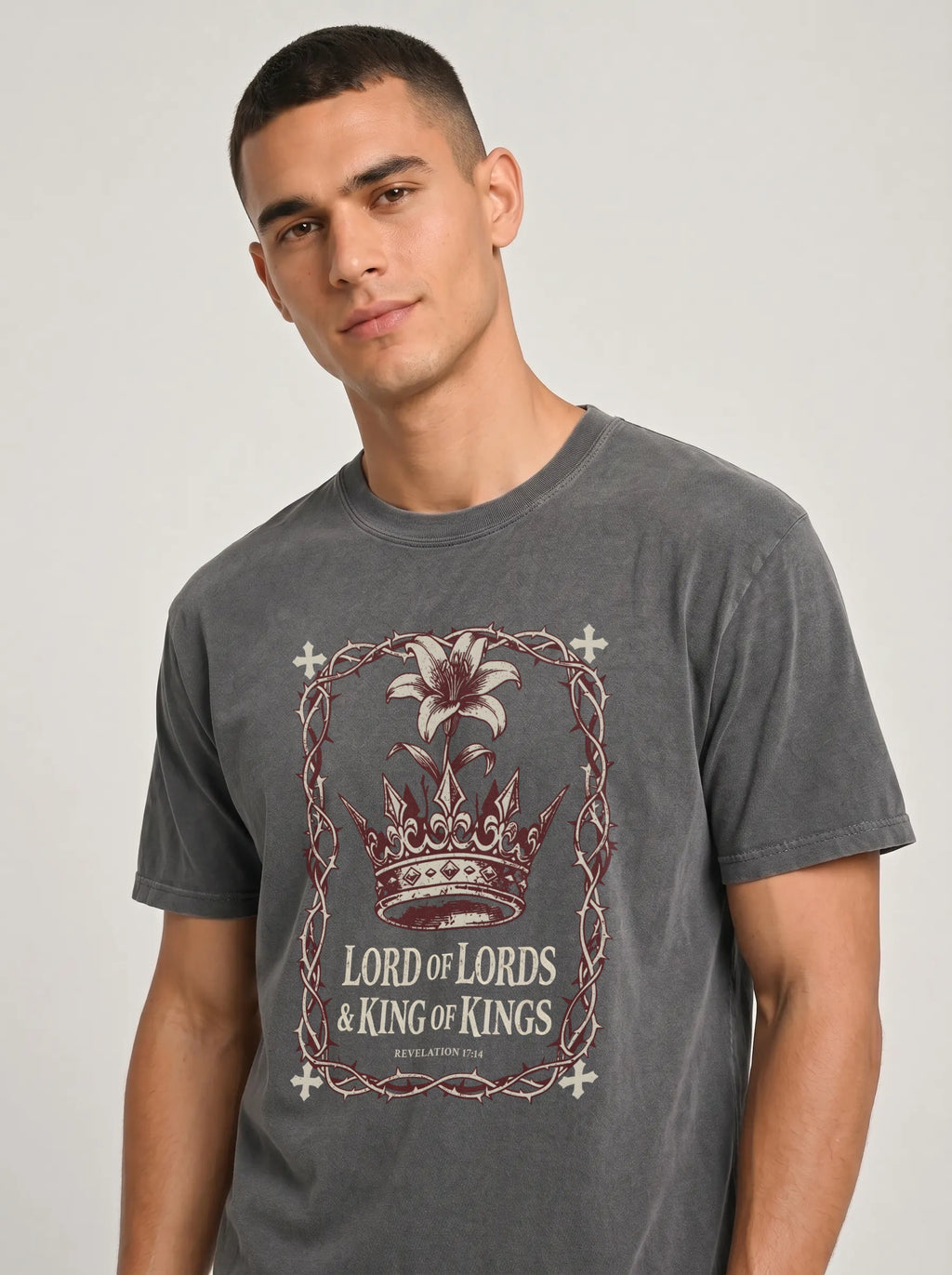 A man wearing a dark gray graphic t-shirt with a crown and 'Lord of Lords & King of Kings' print, photographed against a neutral light gray background.